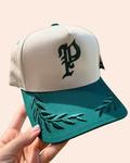 A baseball cap held by a hand, featuring an off-white crown with a dark green embroidered gothic letter 'P' and a dark green brim with embroidered leafy branches.
