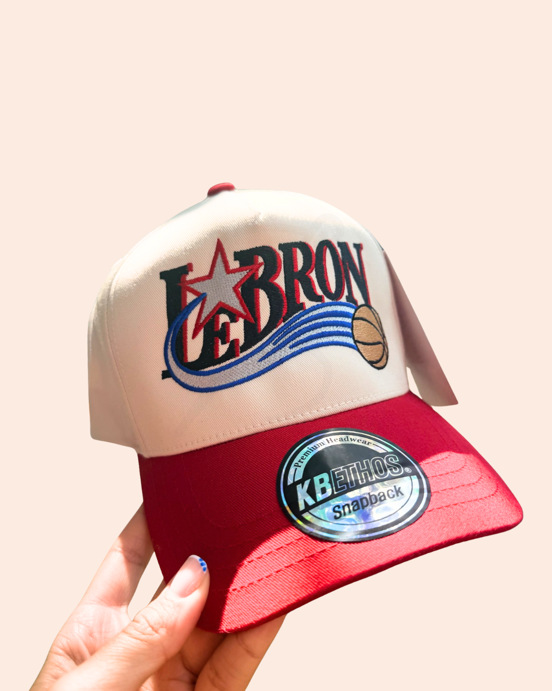A hand holding a snapback cap featuring a cream crown with a large 'LEBRON' graphic and basketball logo, and a red brim.