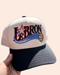 A cream and black trucker style baseball cap featuring a custom 'LEBRON' twist on the Sixers logo embroidered with a red star, blue swoosh, and basketball graphic, held by a hand against a solid pale peach background.