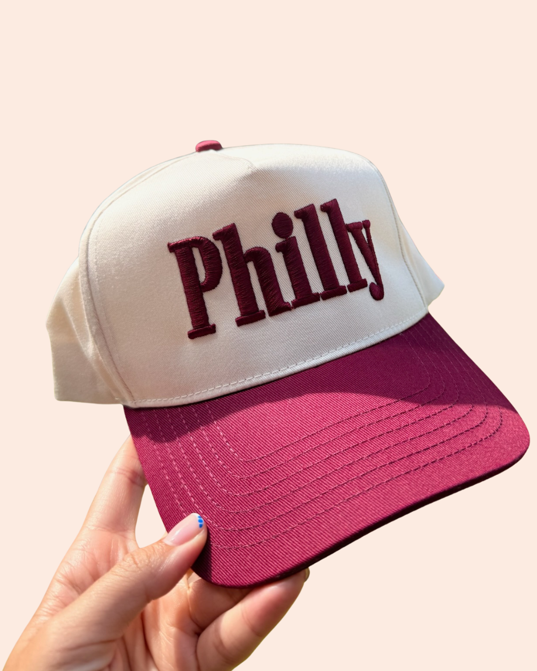 A hand holding a Philadelphia Phillies trucker hat with a cream crown and a maroon brim, featuring the word 'Philly' embroidered in dark red on the front, set against a solid pale peach background.