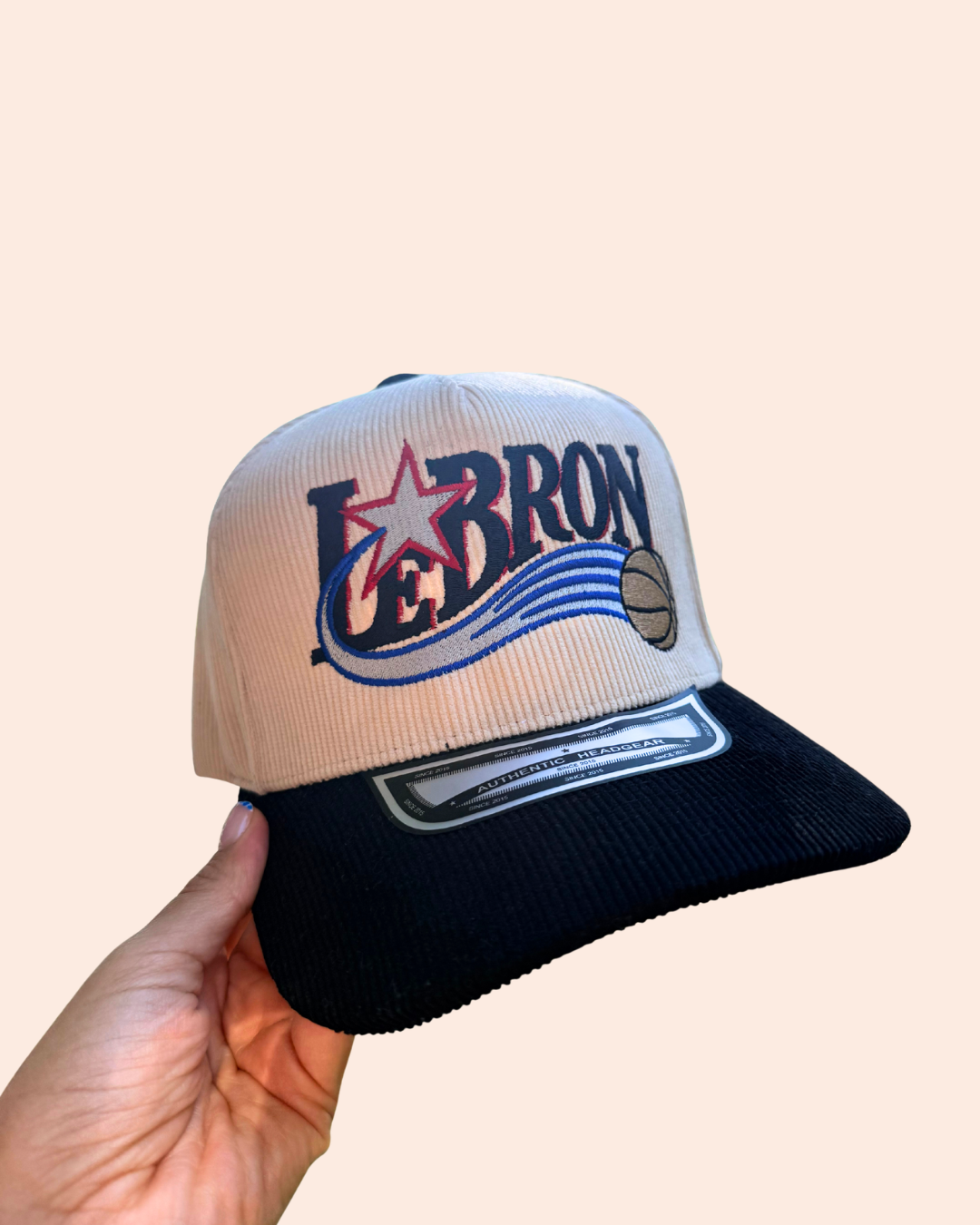 A cream corduroy, trucker style baseball cap held by a hand, featuring a cream-colored crown with a "LeBron" twist on the iconic Sixes logo.