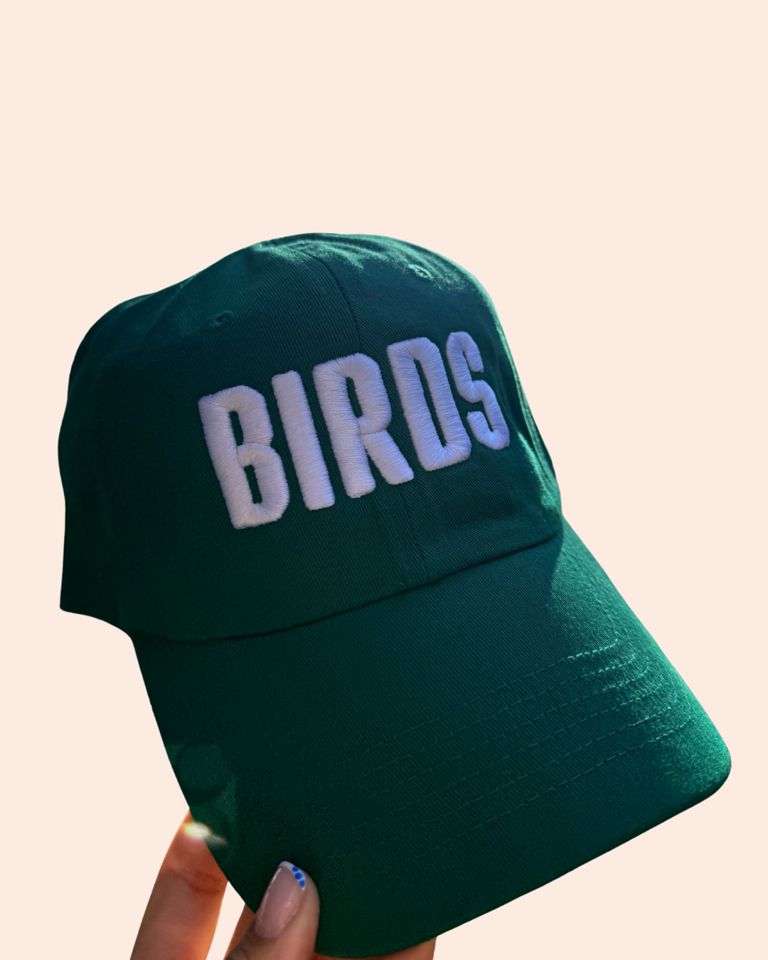 A dark green baseball cap with 'BIRDS' embroidered in white letters on the front, held by a hand against a solid pale peach background.
